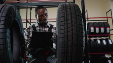 Bir otomotiv mağazasında ya da depoda not alırken tulumlu Afrikalı Amerikalı bir işçi depodaki bir lastiği inceliyor..