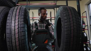 Tulumlu ve eldivenli bir Afrikalı Amerikalı işçi depodaki lastikleri dikkatle inceler ve tamirhanedeki bir müşteri için en iyi seçeneği seçer..