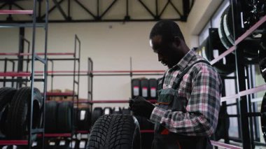 Tulum ve eldiven giyen Afrikalı bir Amerikalı tamirci, bir oto tamirhanesinde lastik izi derinliğini inceler. Müşteriler için güvenlik ve kalite sağlar..