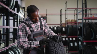 İş tulumlu ve eldivenli bir Afrikalı Amerikalı tamirci mağazadaki bir lastiği incelemek için lastik derinliği göstergesi kullanır. Müşterilerin kalite ve yol güvenliğini sağlar..