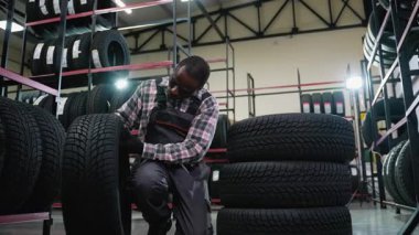 Tulumlu bir tamirci lastik dükkanında diz çöker ve organize bir otomotiv deposunda lastikleri dikkatle inceleyip istifler..