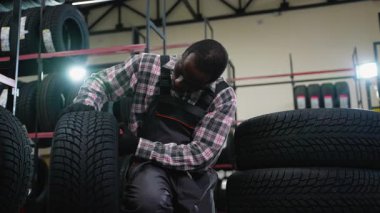Bir otomotiv dükkanında envanter düzenlerken tulumlu bir tamirci lastiği inceliyor. Müşteriler için kaliteli ve düzgün istifleme sağlıyor..
