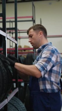 Ekose gömlekli ve tulumlu bir işçi, oto yedek parça mağazasındaki bir müşteri için en iyi seçeneği seçerken raflardaki lastikleri kontrol eder.