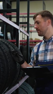 Tulum ve eldiven giyen bir işçi, elinde bir pano tutarken, bir oto yedek parça mağazasında veya depoda envanter kontrolü yaparken, raflardaki lastikleri kontrol eder..