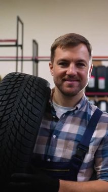 Tulum giymiş neşeli bir işçi, lastik dolu raflarla çevrili bir otomotiv mağazasında ya da depoda omzunda bir lastik tutar..