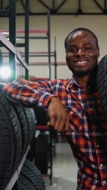 Ekose gömlekli, gülümseyen bir Afrikalı Amerikalı, araba lastikleri ve aksesuarlarla dolu raflarla çevrili bir oto tamirhanesinde lastik taşıyor..
