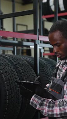 Eldivenli ve üniformalı bir depo çalışanı, otomotiv deposunda bir not panosu ve kalemle envanteri incelerken lastikleri kontrol eder..