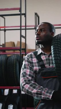 Tulum giymiş neşeli bir işçi, lastik dolu raflarla çevrili bir otomotiv mağazasında ya da depoda omzunda bir lastik tutar..