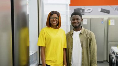 Bir elektronik mağazasında açık bir buzdolabının yanında duran gülümseyen Afrikalı Amerikalı çift, evleri için yeni bir alet seçecekleri için heyecanlılar..
