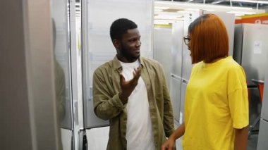 Genç Afrikalı Amerikalı çift, bir mağazada buzdolabının özelliklerini tartışıp tartışırken evleri için en iyi seçeneği seçiyorlar..