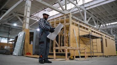 Sert bir şapka ve eldiven takan Hintli bir mühendis, üretim tesisinin içindeki modüler prefabrik evlerin montajını denetlerken planları inceliyor..