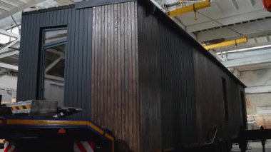 Koyu ahşap kaplamalı prefabrik modüler bir ev güvenli bir şekilde bir fabrikanın içindeki bir nakliye kamyonuna yerleştirilir ve varış yerine ulaştırılmaya hazır hale getirilir..