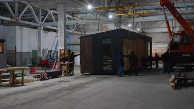Bir grup işçi, modüler bir evi büyük bir sanayi fabrikasının içine taşıyarak, daha fazla montaj veya ulaşım için hazırlıyor..