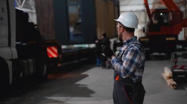 Güvenlik teçhizatlı bir inşaat işçisi modüler bir evi endüstriyel bir depodaki bir kamyona yüklemek için telsiz kullanıyor..