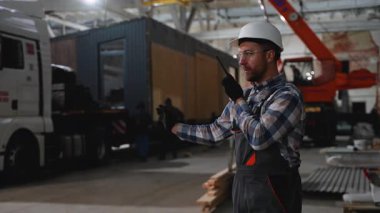 Güvenlik donanımlı bir inşaat işçisi, fabrikadaki bir nakliye kamyonuna prefabrik modüler bir evin yüklenmesini denetlerken radyo aracılığıyla iletişim kuruyor..