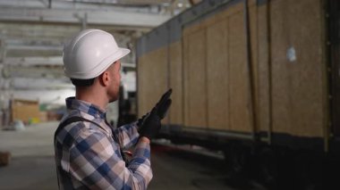 Şapkalı ve eldivenli bir inşaat işçisi modüler bir ev fabrikasındaki SIP panel evin taşınmasını denetliyor..