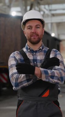 Kasketli ve güvenlik teçhizatlı gülümseyen bir inşaat işçisi modüler bir ev fabrikasında duruyor, lojistik ve prefabrik konut ünitelerinin taşınmasını denetliyor..