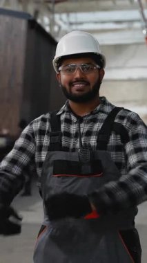 Kasketli ve güvenlik teçhizatlı gülümseyen bir inşaat işçisi modüler bir ev fabrikasında duruyor, lojistik ve prefabrik konut ünitelerinin taşınmasını denetliyor..