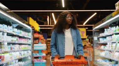 Gözlüklü ve kot gömlekli genç bir kadın, marketin koridorunda, soğutulmuş ürünler ve raflarla çevrili bir alışveriş arabasını itiyor..