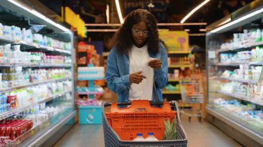Genç Afro-Amerikan kadın gözlüklü ve kot gömlekli süpermarket koridorunda elinde alışveriş arabasıyla dikilirken alışveriş makbuzunu inceliyor..