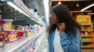 Gözlüklü ve kot gömlekli genç Afrikalı Amerikalı kadın marketin soğutucu reyonundaki yoğurt seçeneklerini dikkatle inceliyor..