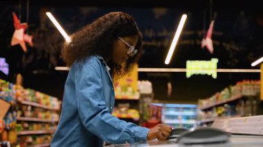 Gözlüklü ve kot gömlekli genç Afrikalı bir kadın dondurucudan donmuş kırmızı balık toplarken dondurulmuş gıda reyonundan alışveriş yapıyor..