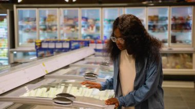 Gözlüklü ve kot gömlekli genç Afrikalı bir kadın süpermarkette dondurucu kapağı açıp dondurulmuş gıda seçeneklerini inceliyor..