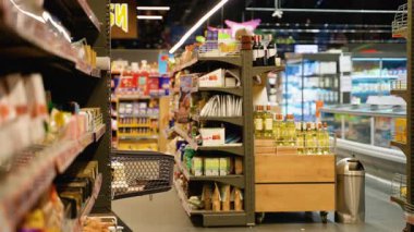 Gözlüklü ve kot gömlekli genç Afrikalı bir kadın bir alışveriş arabasını süpermarket koridoruna itiyor, market ve şarap şişeleriyle dolu rafları karıştırıyor..