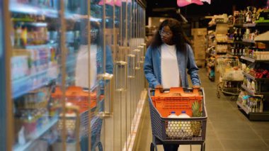 Gözlüklü ve kot gömlekli genç Afrikalı bir kadın bir alışveriş arabasını süpermarket koridoruna itiyor, cam kapılar ardında dondurulmuş ürünlere göz atıyor..
