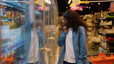 Gözlüklü ve kot gömlekli genç bir kadın süpermarkette soğutucu bölümüne göz atıyor ve cam kapıların arkasından alkolsüz bir içecek seçiyor..