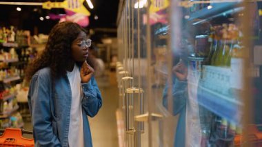 Gözlüklü ve kot gömlekli genç bir kadın süpermarkette soğutucu bölümüne göz atıyor ve cam kapıların arkasından alkolsüz bir içecek seçiyor..
