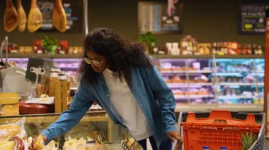 Genç Afro-Amerikalı gözlüklü ve kot gömlekli bir kadın marketten alışveriş yaparken marketten peyniri seçiyor..