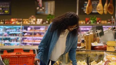 Gözlüklü ve kot gömlekli genç Afrikalı bir kadın süpermarkette tezgahta geziyor, alışveriş yaparken peynir seçiyor..