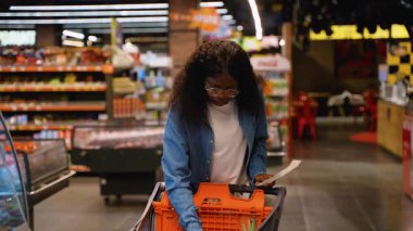 Genç Afrikalı Amerikalı ev hanımı gözlüklü ve kot gömlekli. Market koridorunda alışveriş arabasına bir şeyler yerleştirirken alışveriş makbuzunu inceliyor..