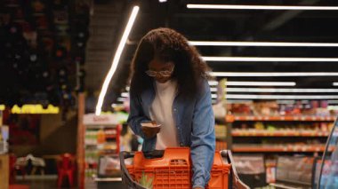 Kot gömlekli genç Afrikalı Amerikalı kadın markette alışveriş yaparken aldığı şok edici ifadeyle alışveriş fişini inceliyor..