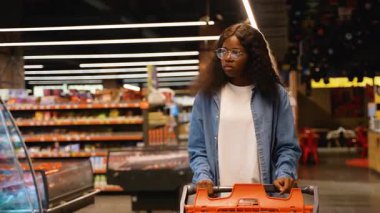 Kot gömlekli genç Afro-Amerikalı bir kadın market koridorunda alışveriş arabasını itiyor, yiyecek ve temel şeyleri seçerken odaklanmış görünüyor..