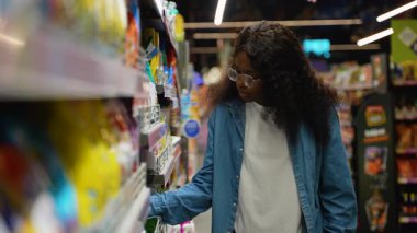 Kot gömlekli genç Afrikalı bir kadın süpermarket koridorunda temizlik malzemesi seçiyor ve farklı deterjan seçeneklerini dikkatlice inceliyor..