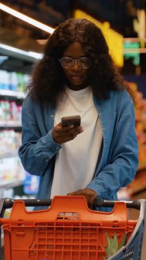 Kot pantolonlu genç Afro-Amerikalı kadın süpermarkette alışveriş yaparken akıllı telefonunu kontrol ediyor, koridorda alışveriş arabasıyla alışveriş yapıyor..