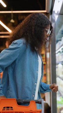 Genç Afro-Amerikalı gözlüklü ve kot gömlekli bir kadın süpermarketin soğutmalı bölümünde süt seçeneklerini incelerken bir yandan da alışveriş arabasını itiyor..