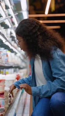Gözlüklü ve kot gömlekli genç Afrikalı bir kadın marketin dondurulmuş süt ürünleri bölümünden yoğurt almak için çömeliyor..