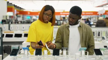 Genç bir Afrikalı Amerikalı çift, bir elektronik mağazasından yeni bir akıllı telefon alıyor. Satın almadan önce özelliklerini, modellerini ve fiyatlarını karşılaştırıyorlar..