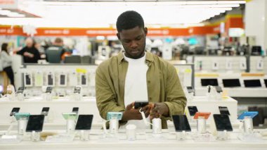 Genç bir Afrikalı Amerikalı, bir teknoloji mağazasında akıllı telefonu inceliyor. Model ve özelliklerini karşılaştırıyor..