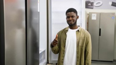 Haki ceketli gülümseyen Afro-Amerikalı bir adam, tercihinden memnun bir şekilde bir buzdolabı seçtikten sonra onay veriyor..
