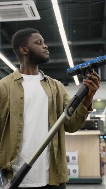Genç Afrikalı Amerikalı bir adam elektrik süpürgesi almak için bir mağazadan alışveriş yapıyor, karar vermeden önce eklerini ve özelliklerini dikkatlice inceliyor..