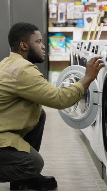 Mutlu bir Afrikalı Amerikalı ev aletleri mağazasında çamaşır makinesini incelemek için çömelir, satın almadan önce davul büyüklüğünü ve özelliklerini kontrol eder..