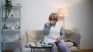 Rahat bir kanepede oturan yaşlı bir kadın kendini kötü hissederken burnunu silmek için mendil kullanıyor. İlaçlar, burun spreyleri ve çay grip veya alerji için evde tedavi öneriyor.