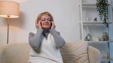 Kıdemli kadın rahat bir koltukta oturuyor, şakaklarını kederli bir ifadeyle tutuyor, muhtemelen baş ağrısı, migren ya da gerginlikle ilgili rahatsızlığı var..