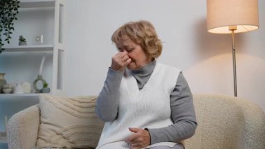 Yaşlı bir kadın rahat bir koltukta oturuyor, burnunu acı dolu bir ifadeyle tutuyor, muhtemelen sinüs basıncı, burun tıkanıklığı ya da hastalıktan dolayı baş ağrısı çekiyor..