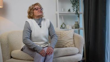 Yaşlı bir kadın rahat bir koltukta oturuyor, dizini tutarak acı çekiyor, muhtemelen eklem ağrısı, eklem iltihabı ya da yaşlanma nedeniyle hareket sorunu yaşıyor..