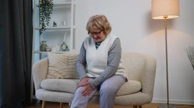 Rahat bir kanepede oturan yaşlı bir kadın, dizini acıdan tutuyor. Yaşlanma nedeniyle eklem ağrısı, eklem iltihabı ya da hareket sorunu yaşıyor gibi görünüyor..